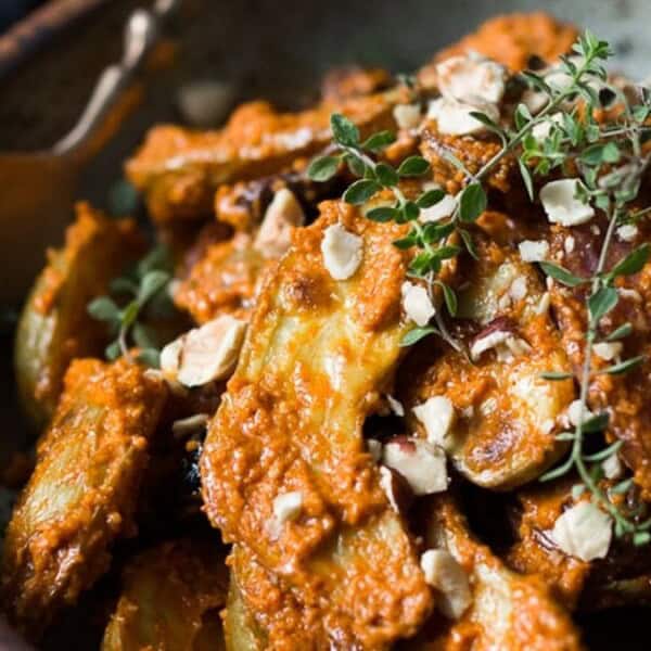 The image features a bowl of roasted vegetables, including potatoes, topped with a red sauce and garnished with chopped nuts and fresh herbs. A spoon is placed inside the bowl.