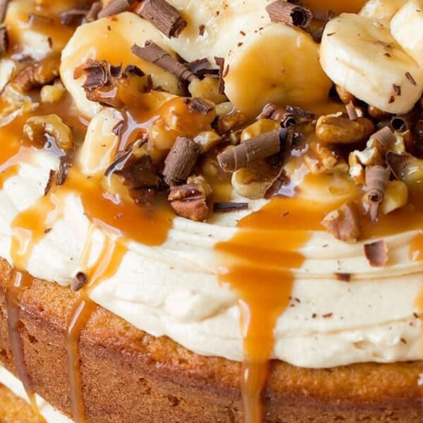 The image shows a layered cake topped with bananas, chocolate shavings, nuts, and a caramel drizzle.
