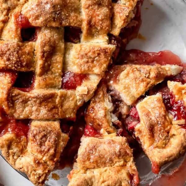 A lattice-topped strawberry rhubarb pie with a slice removed sits on a glass pie dish. A serving utensil and fork are placed nearby on the marble surface.