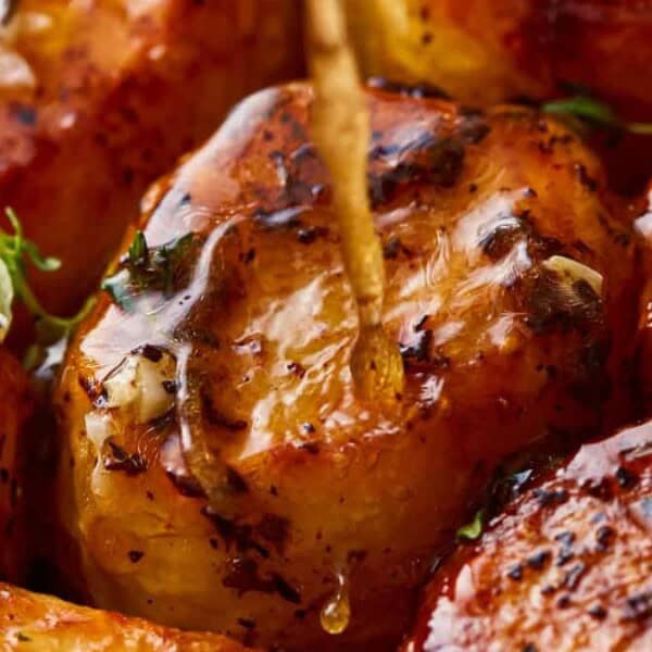 The image displays seared scallops with a golden-brown caramelized surface. They are garnished with herbs and garlic, and one scallop has a toothpick inserted into it.
