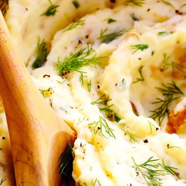 In the image, there is a bowl filled with creamy mashed potatoes. The dish is garnished with fresh dill and sprinkled with black pepper. A wooden spoon is placed in the bowl.