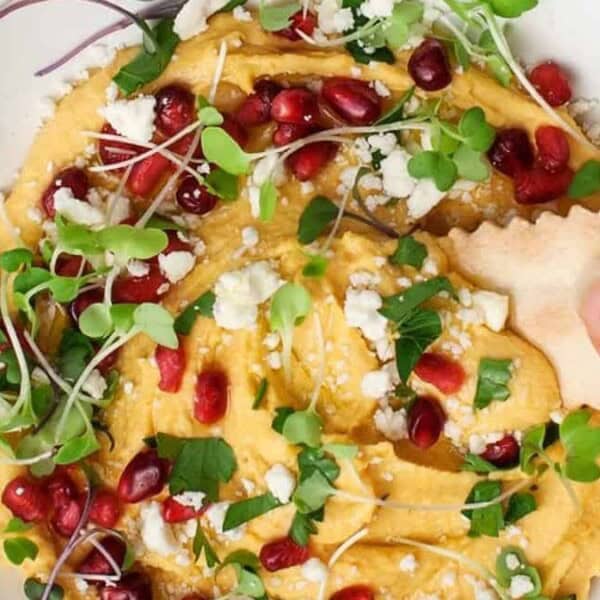 A bowl of hummus is topped with microgreens, pomegranate seeds, and crumbled cheese. A hand dips a cracker into the hummus.
