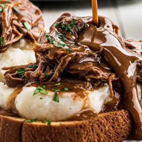 An open-faced sandwich on toasted bread features mashed potatoes and shredded beef, topped with brown gravy and garnished with herbs.
