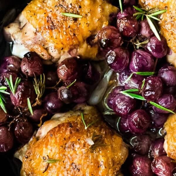The image shows a black skillet containing roasted chicken thighs with crispy skin, surrounded by red grapes and garnished with sprigs of rosemary.