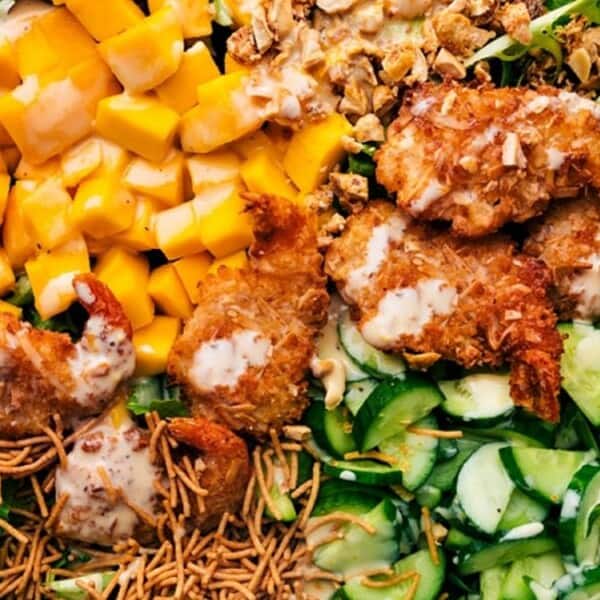 The image features a white plate containing mango, fried chicken pieces, cucumber slices, crunchy noodles, and walnuts arranged as a salad.