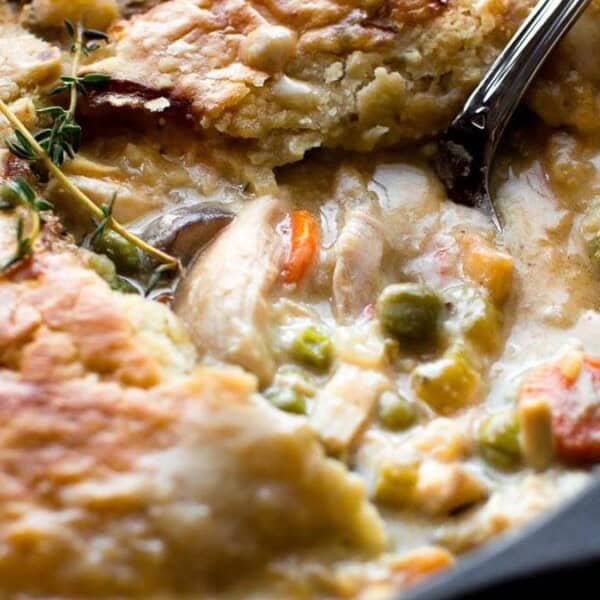 The image shows a pot pie in a black skillet with a golden crust. The creamy filling contains vegetables such as peas and carrots. A sprig of thyme is on the crust, and a spoon is placed in the filling.