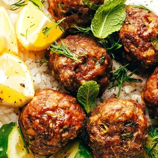 A plate featuring meatballs, lemon wedges, mint leaves, cucumber slices, and rice.