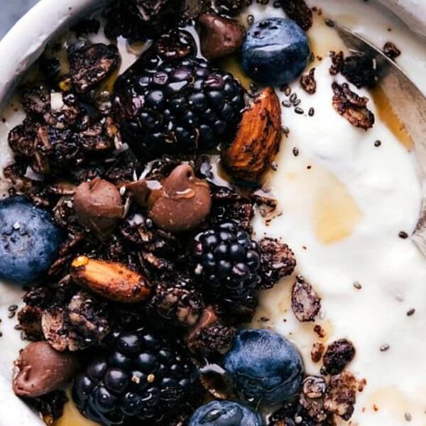 A bowl of yogurt topped with fresh blackberries, blueberries, nuts, chocolate chunks, and granola. A spoon rests in the bowl, and the background is a soft gray surface.