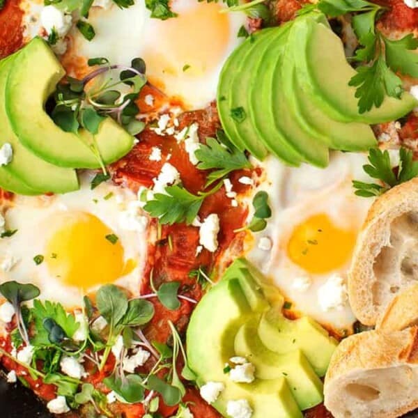 A skillet with shakshuka topped with sunny-side-up eggs, sliced avocado, and crumbled cheese. Fresh cilantro and microgreens are sprinkled on top. Slices of bread are placed to the side.