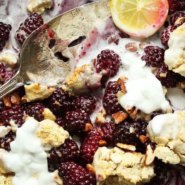 A close-up of a blackberry crumble in a skillet. The dessert features blackberries, chunks of crumble topping, and dollops of whipped cream. A spoon rests in the pan next to a slice of lemon.