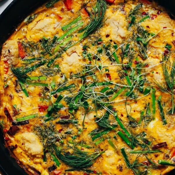 The image shows a frittata in a skillet, topped with asparagus, herbs, and melted cheese. Nearby, there is a small wooden spoon holding grated cheese and part of a yellow cloth can be seen.