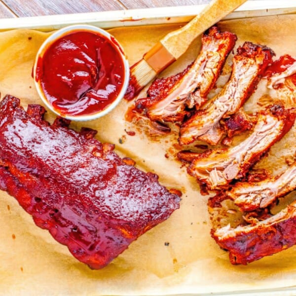 A tray with BBQ ribs, some whole and others sliced, is arranged. Nearby on parchment paper are a cup of barbecue sauce and a brush.