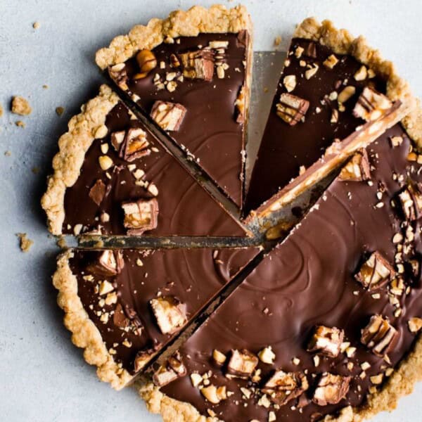 The image displays a chocolate tart with a crumbly crust, topped with pieces of chocolate and nuts. It is sliced into six pieces and placed on a gray surface.