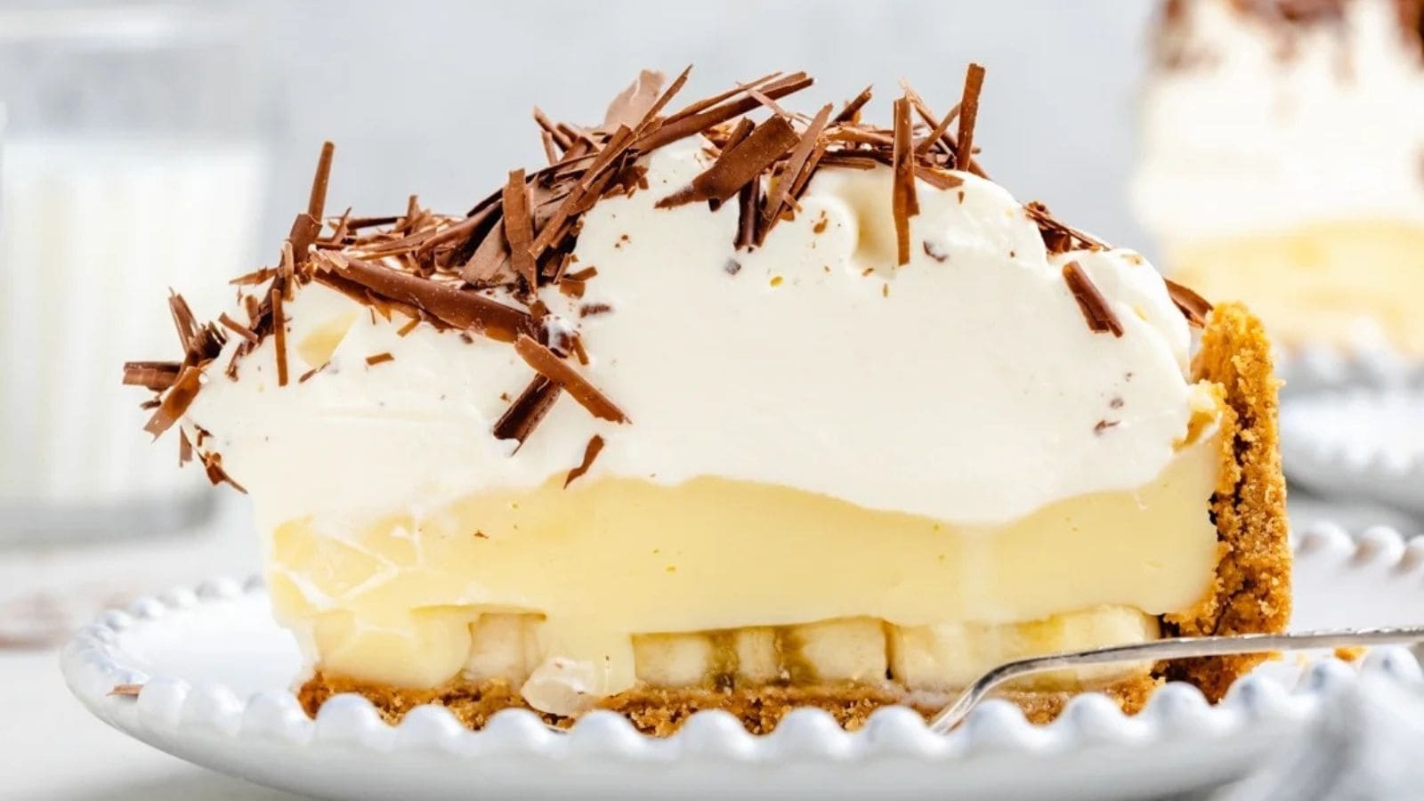 A slice of banana cream pie with a graham cracker crust, topped with whipped cream and chocolate shavings on a white plate.