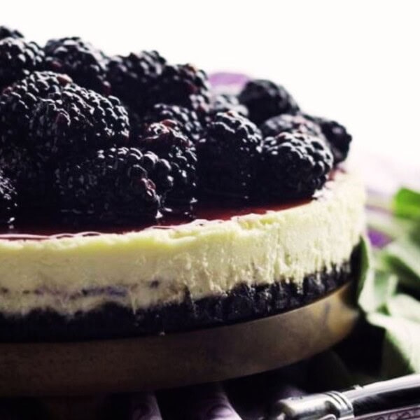 A cheesecake topped with glossy blackberries on a wooden serving board. The creamy filling sits on a dark crust, with a shiny berry glaze. A metal utensil lies beside the cake, and green leaves partially frame the image.