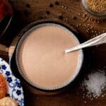 A bowl of creamy sauce with a spoon, surrounded by shrimp, a jar of paprika, mustard seeds, and coarse salt on a wooden surface.