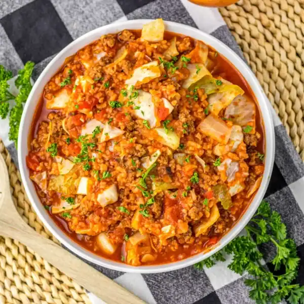 A bowl of soup containing ground meat, tomatoes, and chopped cabbage, garnished with parsley, sits on a checkered cloth next to a wooden spoon and a woven placemat.
