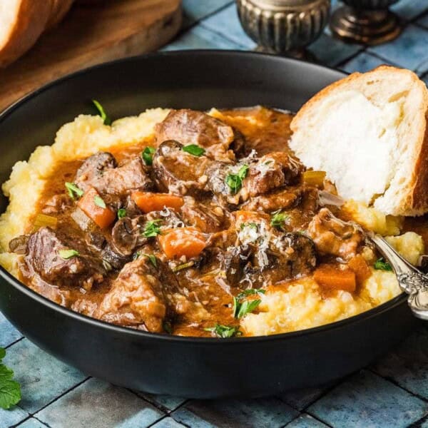 A bowl of savory beef stew with vegetables over creamy polenta, garnished with herbs, beside a slice of crusty bread.