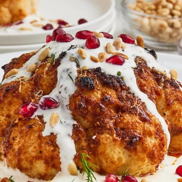 A roasted cauliflower head topped with yogurt sauce, pomegranate seeds, and pine nuts, garnished with fresh herbs on a white plate.