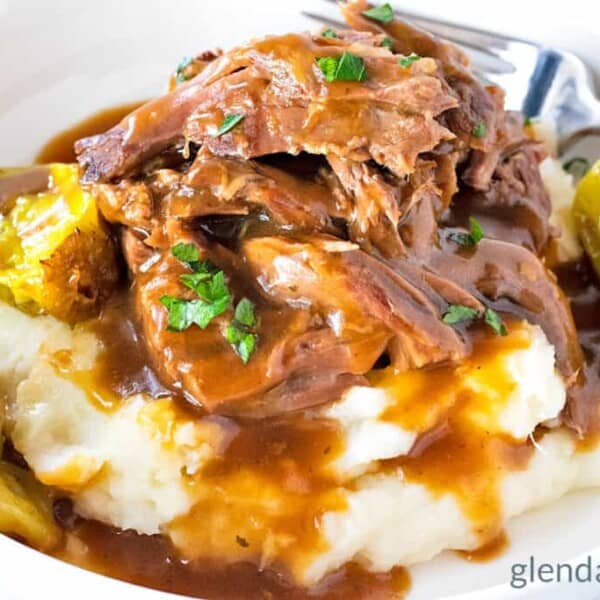 Shredded pot roast with brown gravy and pepperoncini peppers, served over mashed potatoes in a white bowl, with a fork.