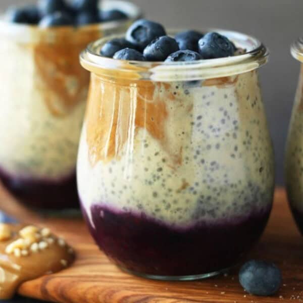 The image shows three glass jars containing layers of chia pudding, blueberry puree, and peanut butter. Each jar is topped with fresh blueberries and placed on a wooden board.