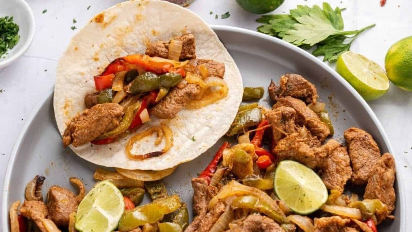 Grilled chicken strips, sautéed onions, bell peppers, lime wedges, and a flour tortilla arranged on a plate.