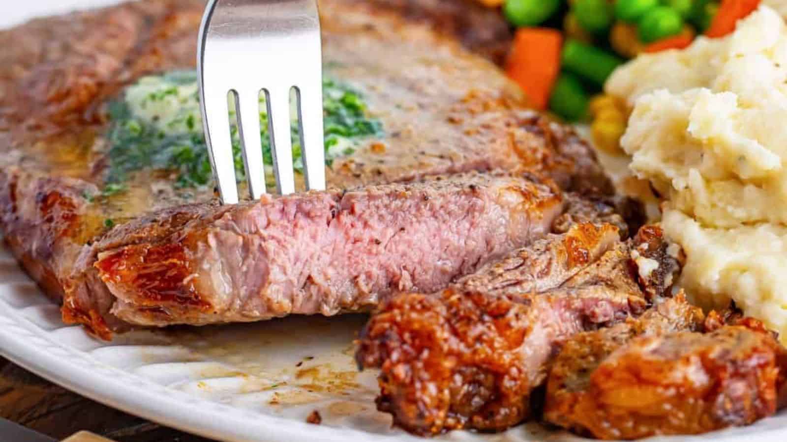 A plate with a medium-cooked steak topped with herb butter, mashed potatoes, mixed vegetables, and a fork cutting into the steak.