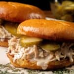 Two sandwiches with chopped chicken, creamy sauce, and pickle slices on toasted buns, arranged on a patterned plate.