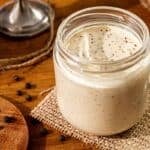 A glass jar filled with a creamy white sauce, topped with ground black pepper, sits on a piece of burlap beside a metal spoon and scattered peppercorns on a rustic wooden table.