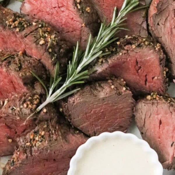Slices of cooked beef tenderloin arranged on a plate, accompanied by a sprig of rosemary and a small dish of white sauce.