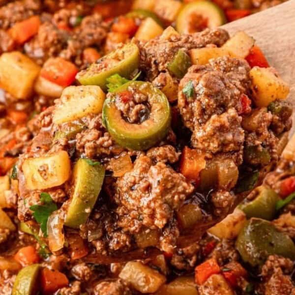 A wooden spoon holds seasoned ground beef, diced potatoes, green olives, and vegetables coated in a thick sauce.