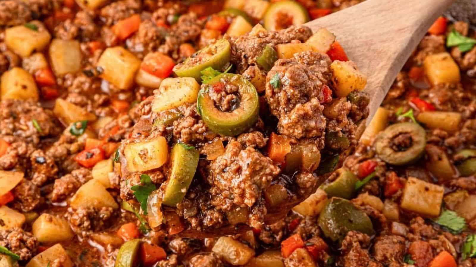 A wooden spoon holds seasoned ground beef, diced potatoes, green olives, and vegetables coated in a thick sauce.