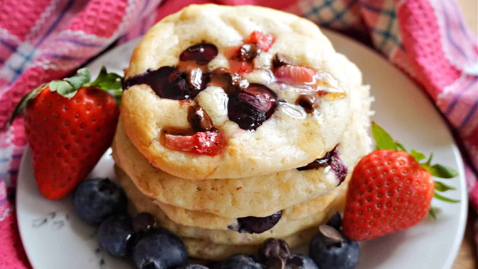 A stack of pancakes topped with blueberries, chocolate chips, and fresh strawberries, served on a white plate.