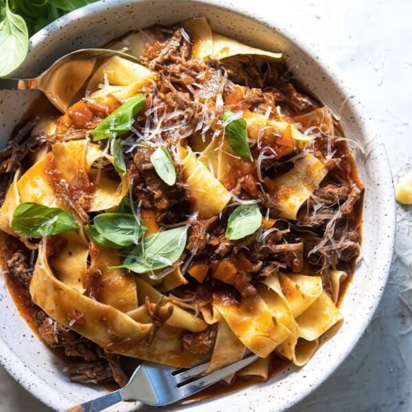 Bowl of pappardelle pasta with beef ragu, tomato sauce, grated cheese, basil leaves; fork and spoon beside the bowl.