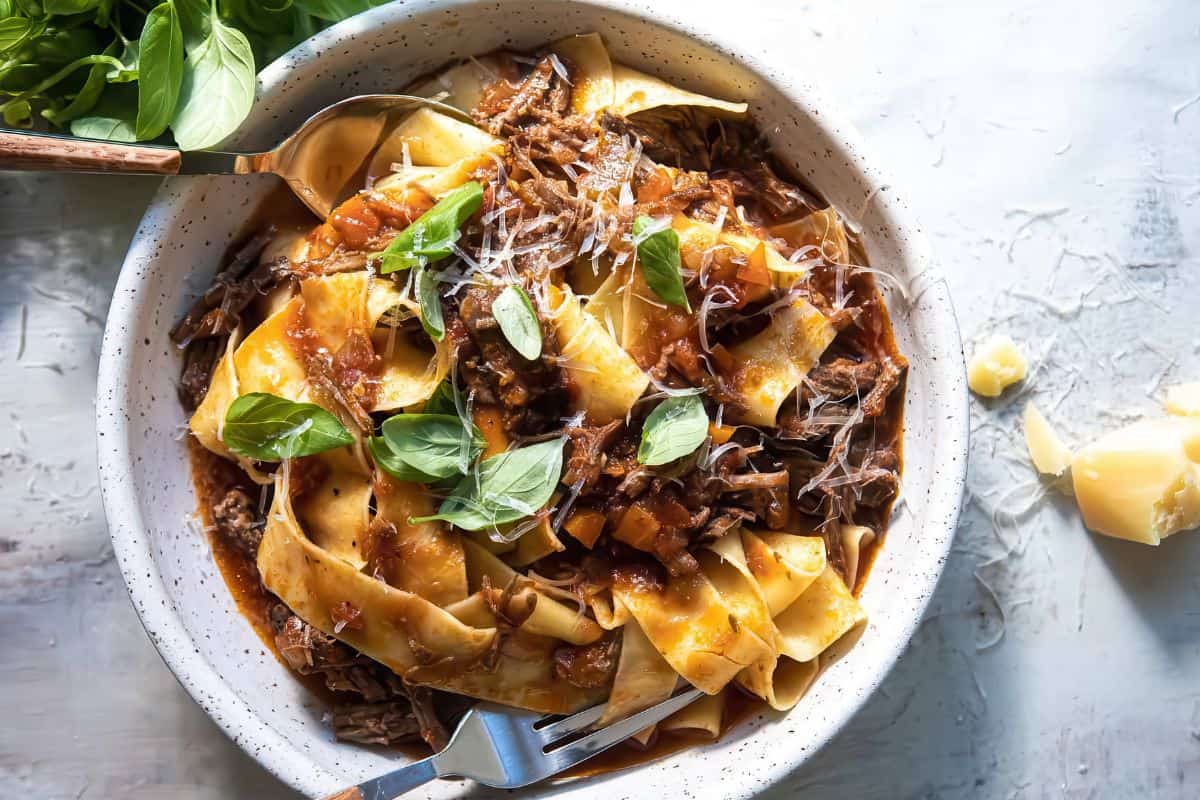A bowl of wide pappardelle pasta topped with shredded beef ragu, tomato sauce, grated cheese, and fresh basil leaves. A fork and spoon are placed in the bowl on a light surface.