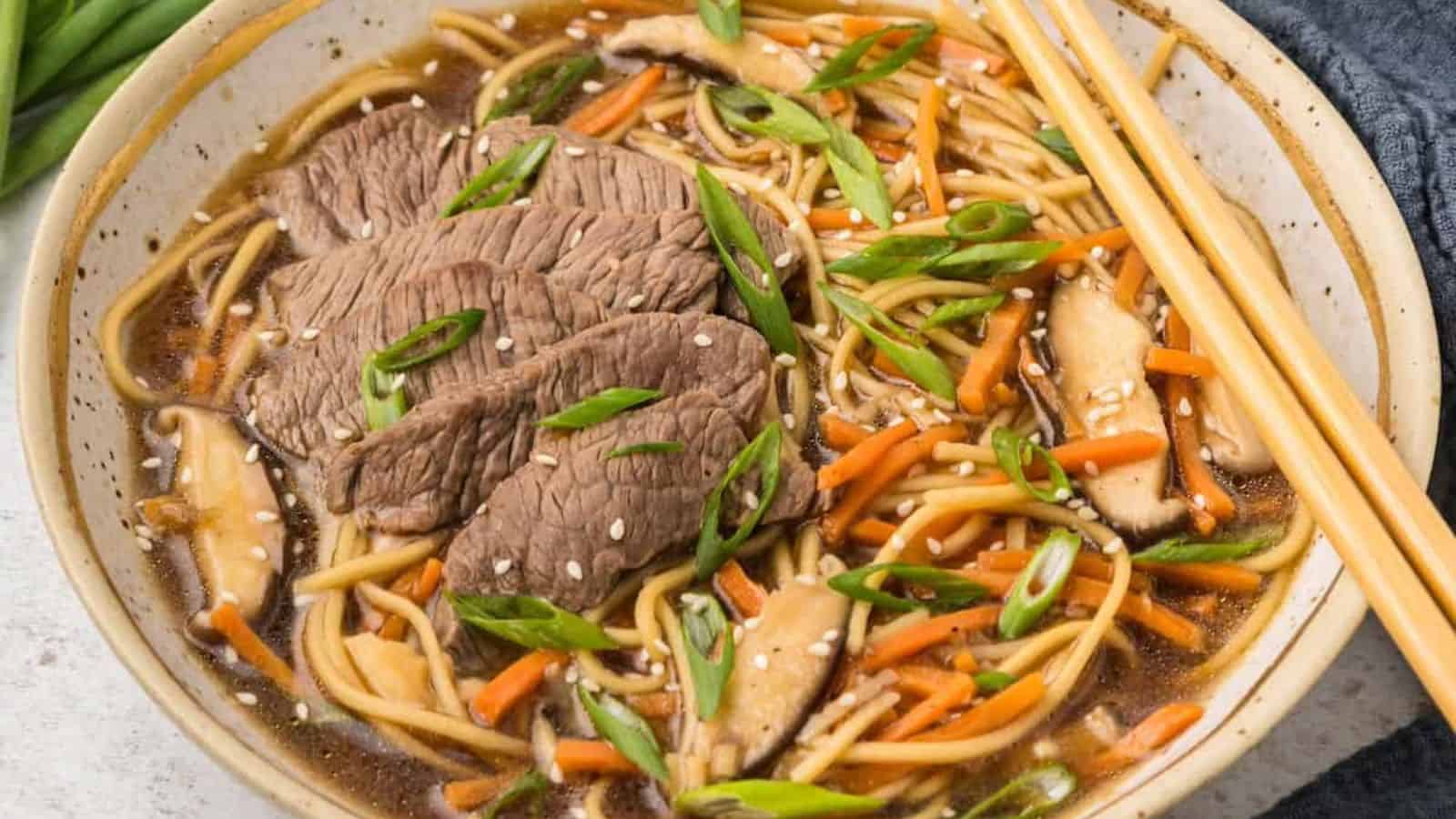 A bowl of noodle soup containing sliced beef, carrot slices, mushrooms, green onions, and sesame seeds, with chopsticks placed on top of the bowl.