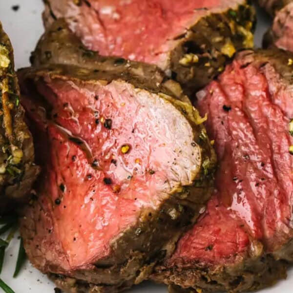 Slices of medium-rare beef tenderloin with a seasoned crust are arranged on a white plate and garnished with rosemary.