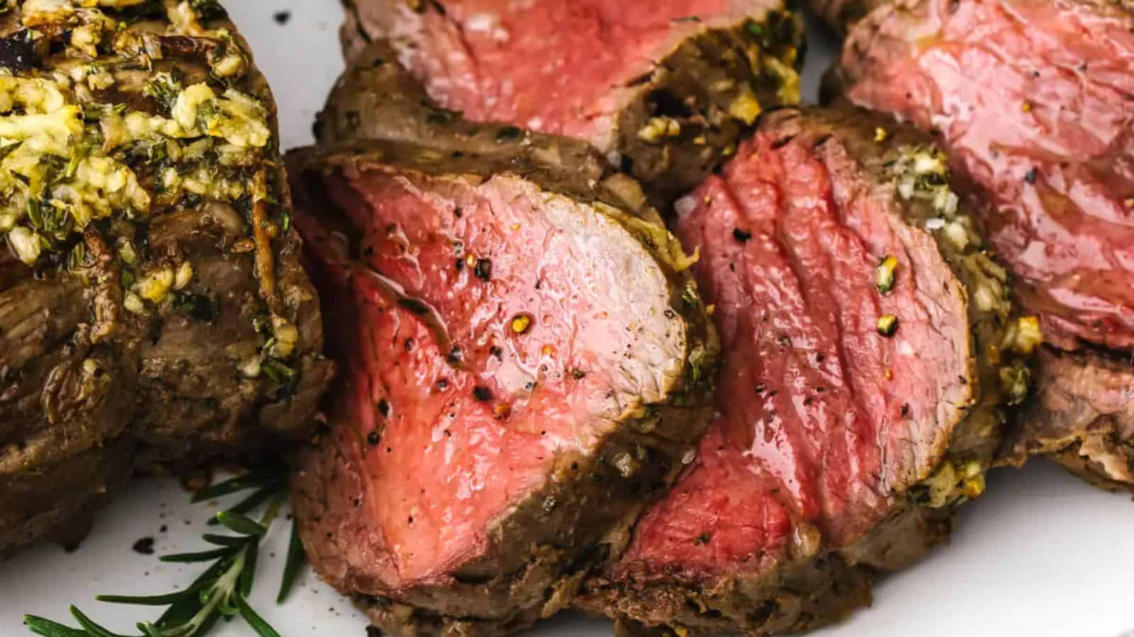 Slices of medium-rare beef tenderloin with a seasoned crust are arranged on a white plate and garnished with rosemary.