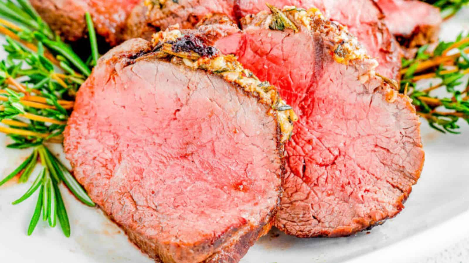 Slices of medium-rare beef tenderloin topped with sprigs of fresh rosemary on a white plate.