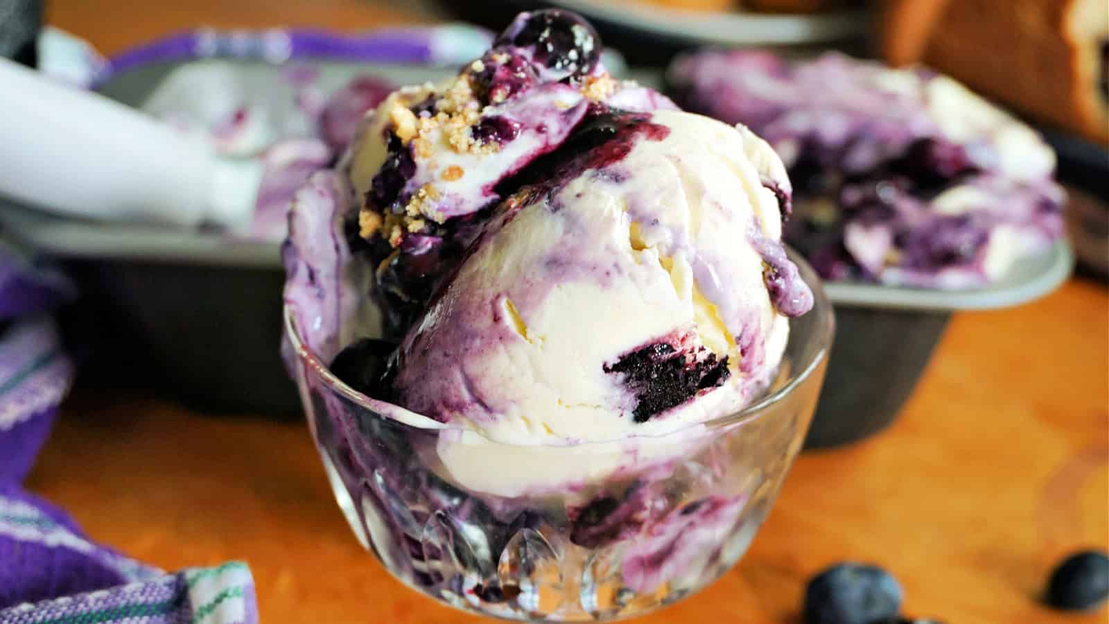 A glass bowl of blueberry swirl ice cream topped with blueberry sauce and crumbs, with an ice cream pan and scoop in the background.
