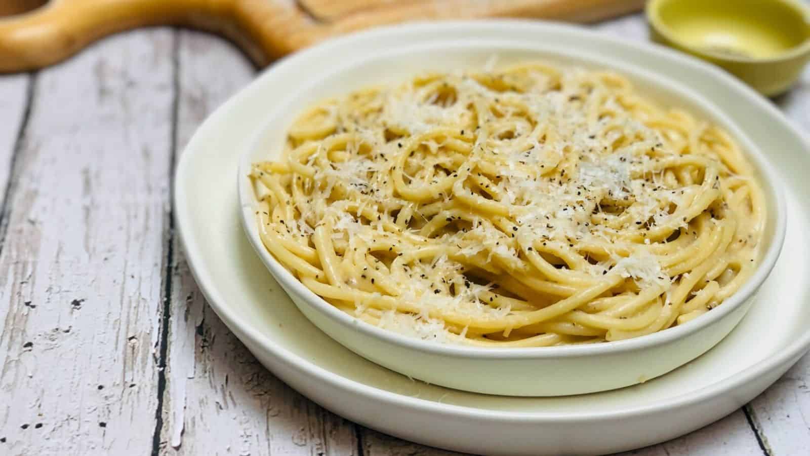 A bowl of spaghetti topped with grated cheese and black pepper, set on a white plate resting on a rustic wooden table.