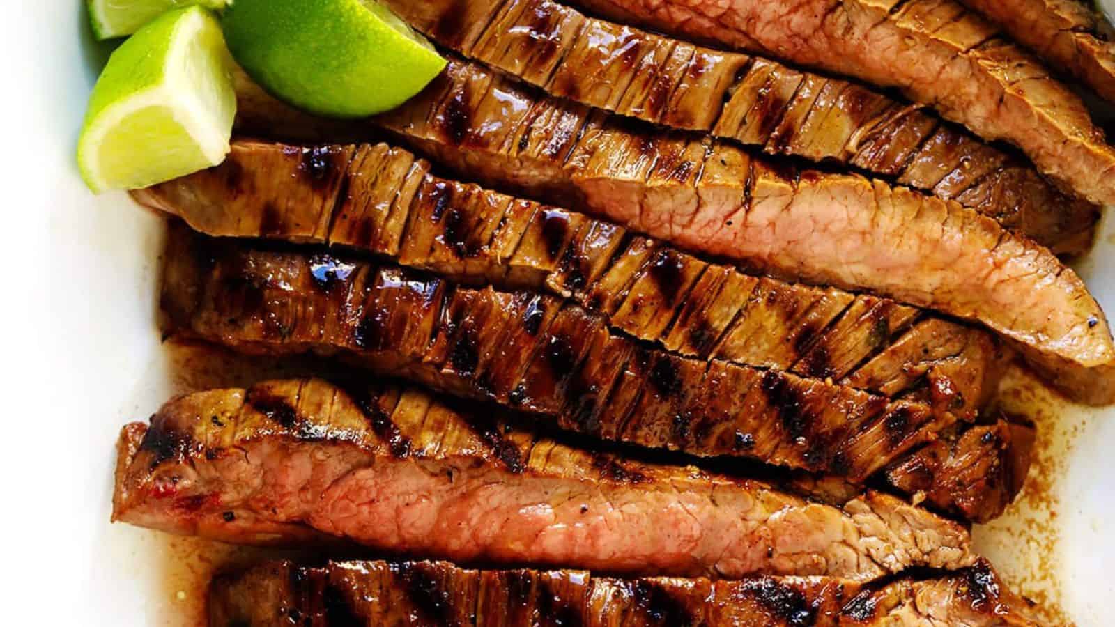 Sliced grilled steak with visible grill marks, arranged on a plate with lime wedges beside it.