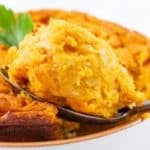 A spoon holds a portion of golden-brown cornbread casserole from a dish, garnished with a parsley sprig, showing its fluffy texture beneath the surface.