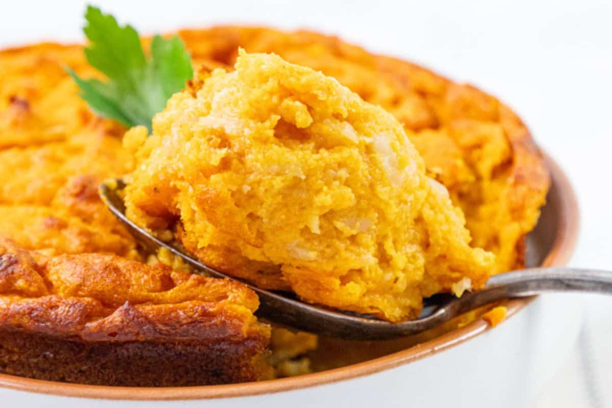 A spoon holds a portion of golden-brown cornbread casserole from a dish, garnished with a parsley sprig, showing its fluffy texture beneath the surface.