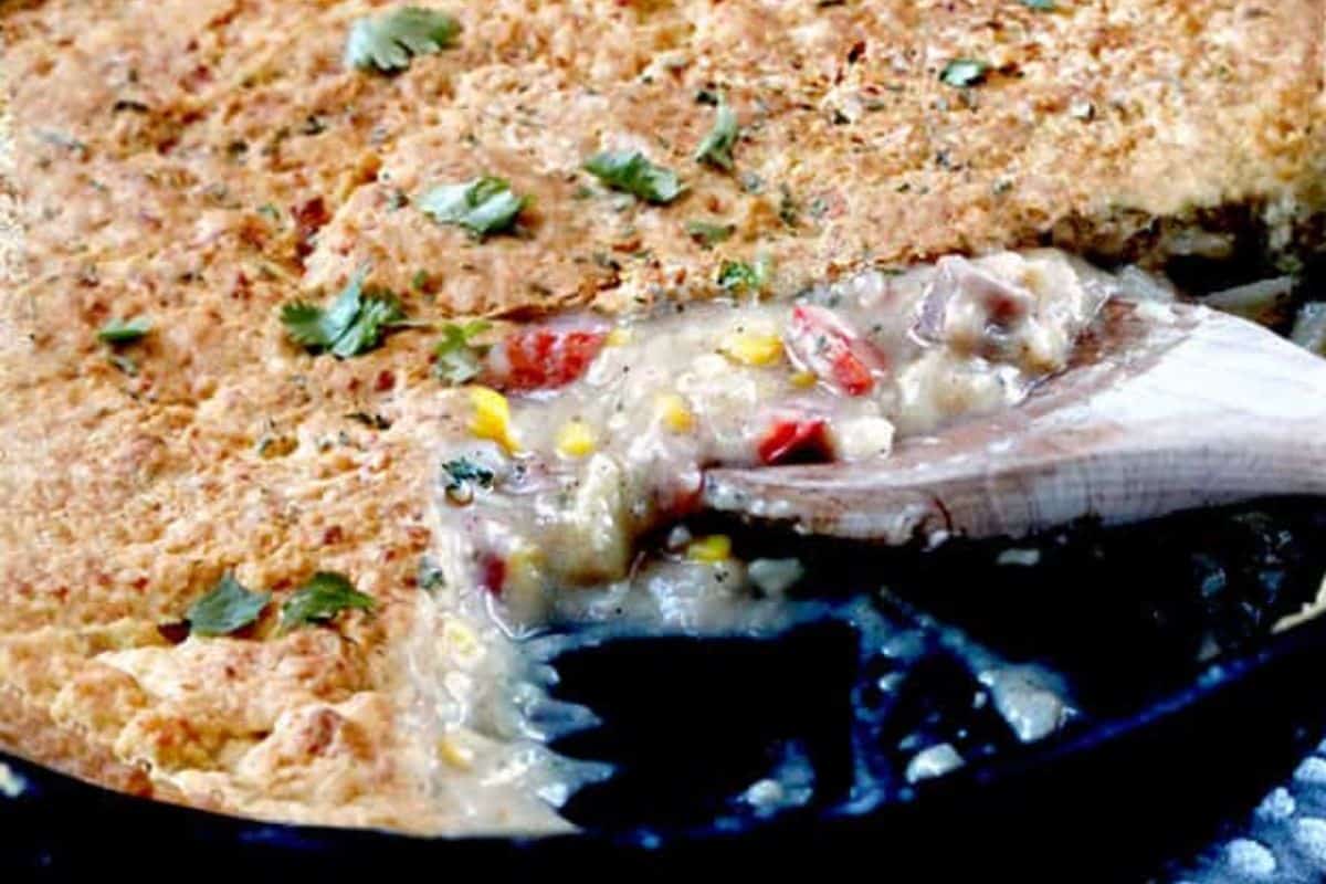 A close-up of a skillet filled with creamy pot pie topped with a golden, flaky crust. A wooden spoon scoops out the filling, revealing vegetables like corn and peppers. Fresh herbs are sprinkled on top.