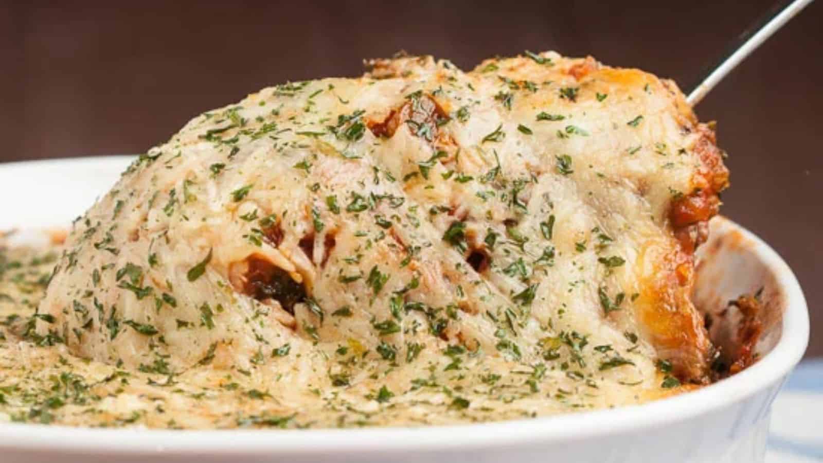 A portion of baked pasta with melted cheese and herbs is being lifted from a white baking dish.