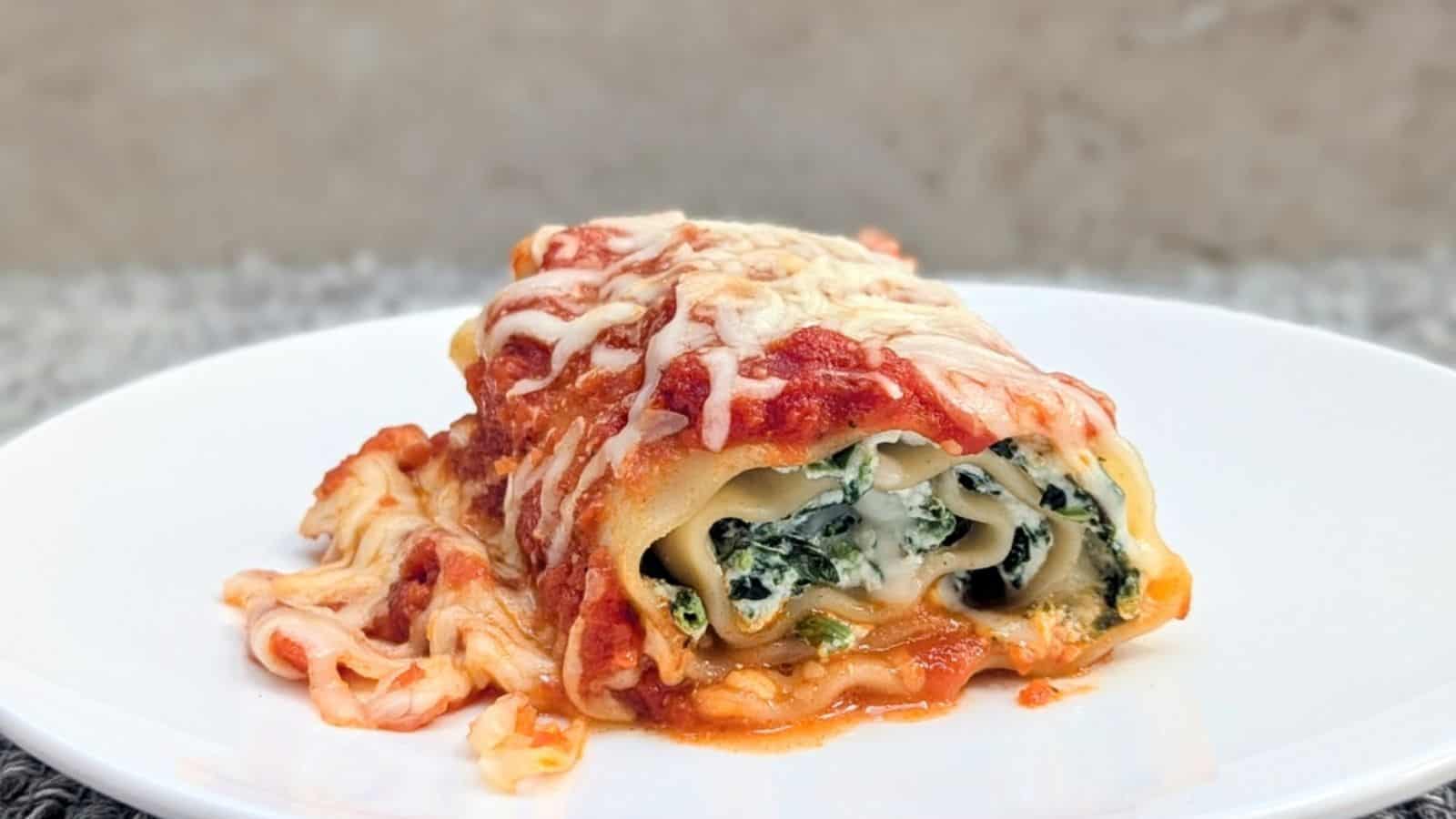 A single spinach and ricotta lasagna roll-up sits on a white plate, topped with marinara sauce and melted cheese.