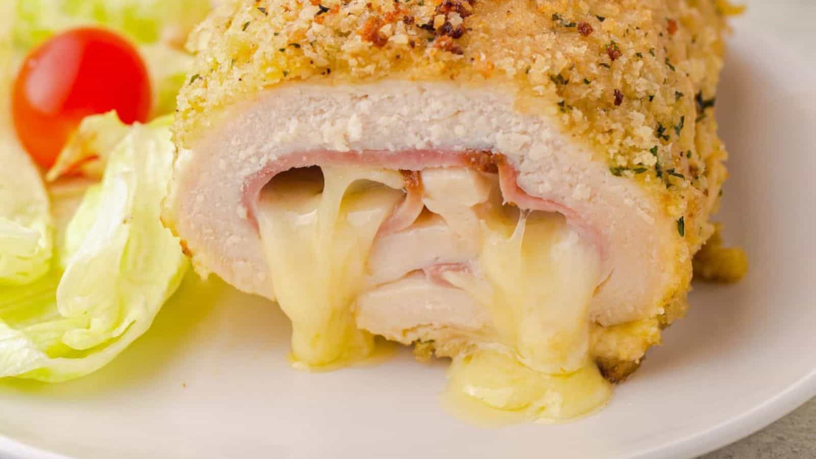 A breaded cutlet filled with ham and melted cheese, accompanied by lettuce and a cherry tomato on a white plate.