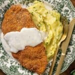 A plate with chicken fried chicken covered in white gravy and a side of mashed potatoes sprinkled with herbs. A golden fork and knife are placed beside the plate.