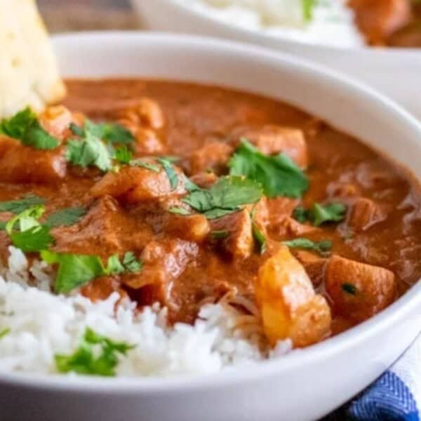 A white bowl containing white rice and chicken curry topped with chopped cilantro, with naan bread on the side. Another similar bowl appears in the background.